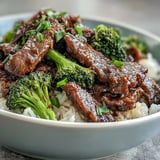 Sliced green onions and sesame seeds garnish a fresh serving of Beef and Broccoli Bowl, steaming beside chopsticks.