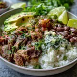 A vibrant carnitas bowl topped with creamy avocado slices, cilantro, and lime wedges.